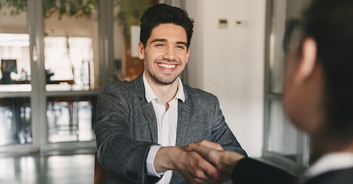 Smiling man in a gray blazer shakes hands with a colleague in an office setting.