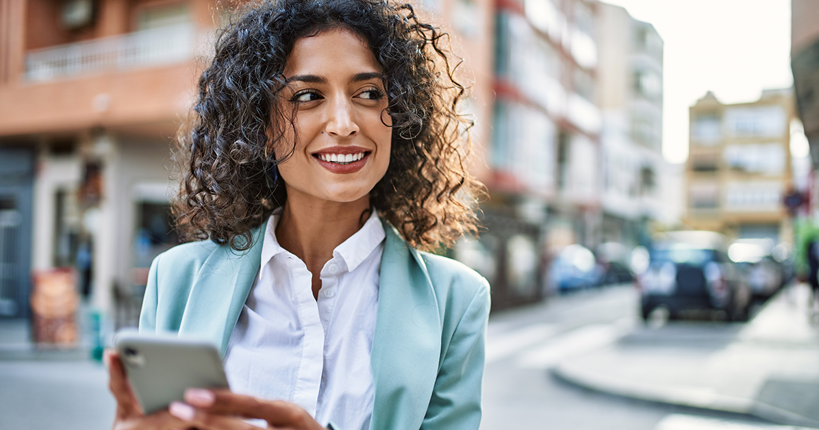 Smiling woman in a light blue blazer holds a smartphone on a sunny urban street.