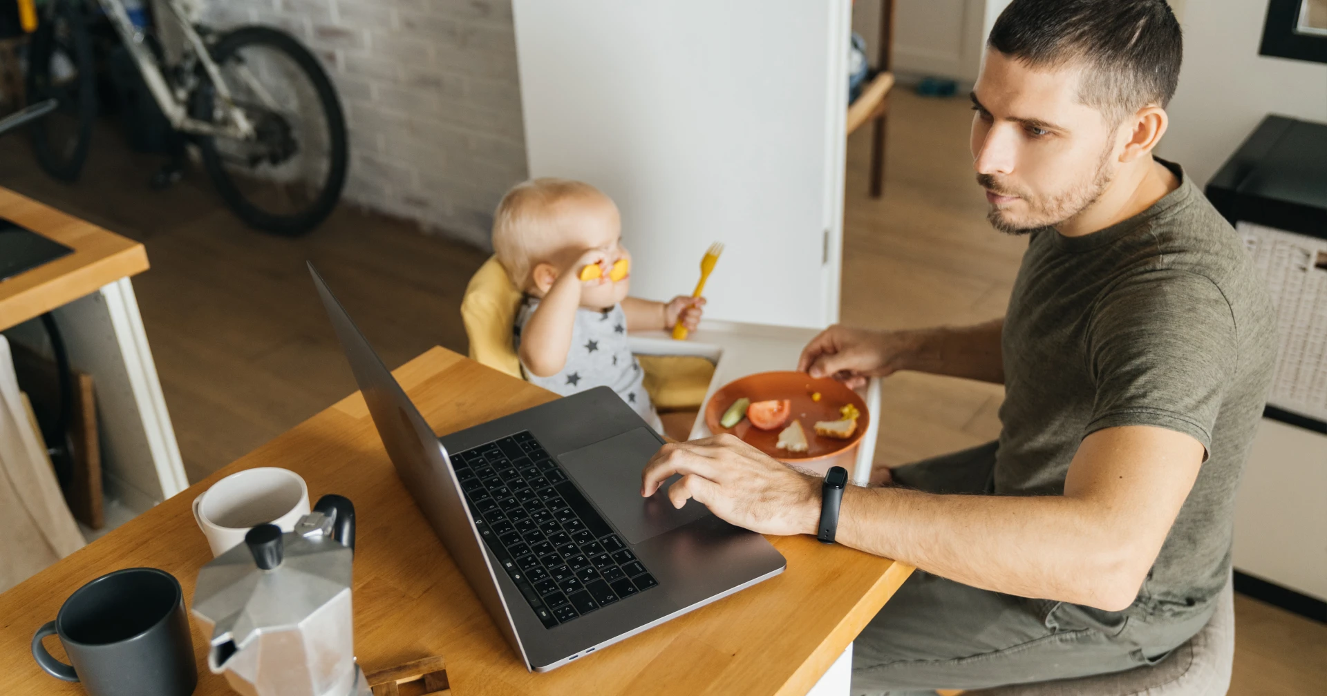 Adult man at a kitchen table using a laptop; baby in a high chair eats with a yellow spoon on a plate nearby.