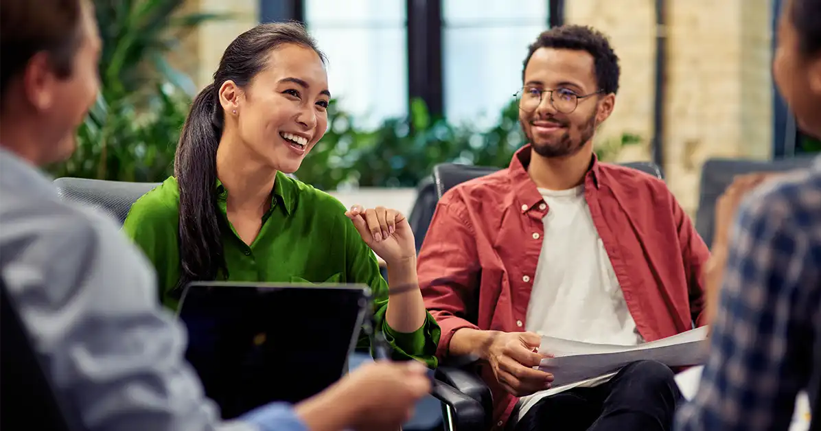 Group of ethically diverse coworkers sitting together talking. There is an Asian woman in a green shirt and an African man next to her in a white shirt with a pink button-up. Blog title: How To Increase Diversity In The Workplace The Right Way