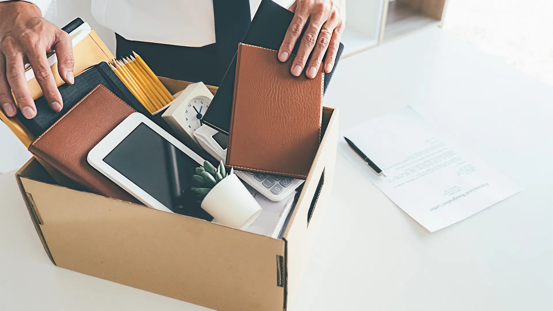 Person reaching into a cardboard box filled with office items: wallets, a tablet, clock, calculator, envelopes, notebooks, and a small plant nearby