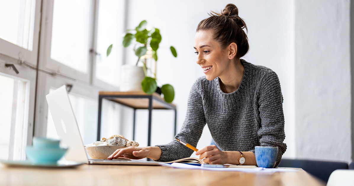 Woman working on a laptop at a sunlit desk with a plate of croissants, notebook, pencil, and a blue mug nearby.