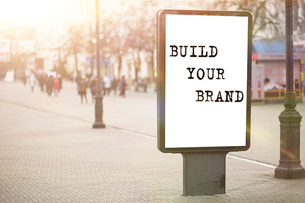 Outdoor street advertisement: a pedestrian area with a freestanding sign that reads 'BUILD YOUR BRAND' in bold letters under warm evening light, suggesting brand-building.