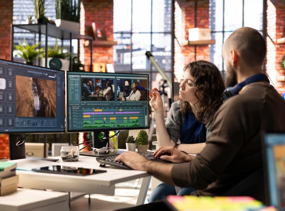 Two creative professionals collaborating on video editing software using dual computer monitors in a modern studio workspace.